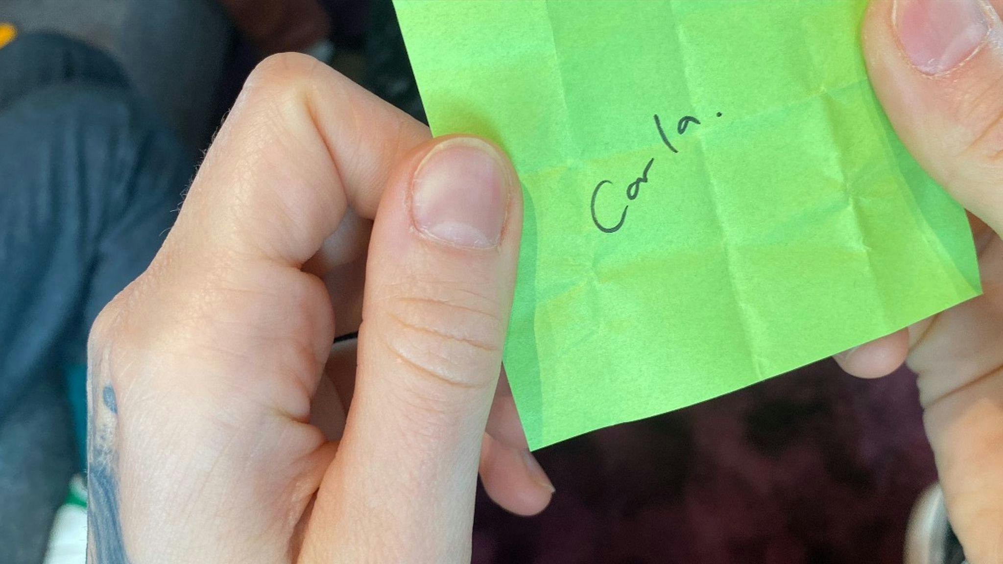 A hand holding a green post-it with the name "Carla" written on it.